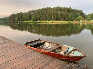 Obraz premium Wooden boat on the pier on the background of the lake. Fishing boat on the shore of a forest reservoir. The boat is tied to the shore. Concept: outdoor recreation, tranquility by the water.