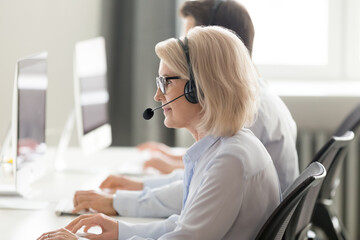 Mature female call center operator in wireless headset consulting client online, working on...