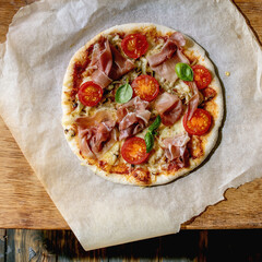 Fresh baking homemade pizza napolitana with prosciutto ham, cheese, tomatoes, basil on baking paper over wooden table background. Home baking or delivered fast food. Flat lay, space.