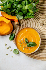 Bowl of pumpkin or carrot vegetarian cream soup decorated by fresh basil, olive oil and pumpkin seeds on white texture background with ingredients above. Flat lay, copy space