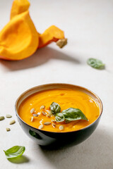 Bowl of pumpkin or carrot vegetarian cream soup decorated by fresh basil, olive oil and pumpkin seeds on white texture table with ingredients above.