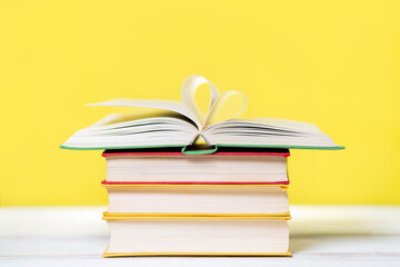 A stack of books with an open book at the top on a yellow background. The pages are wrapped in a heart shape. Copy space. Concept of education and reading