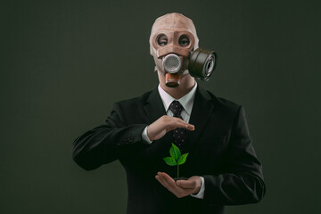 Portrait of a man wearing a business suit and an old gas mask while protectively holding a green plant, isolated on dark green background.