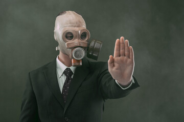Portrait of a man wearing a dark business suit and a gas mask showing his left palm, isolated on dark green background with fog.