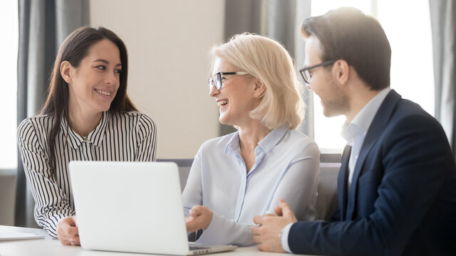 Happy employees interns discussing new corporate software, having fun, pleasant conversation, using laptop, mature coach mentor training staff, explaining online project, horizontal photo
