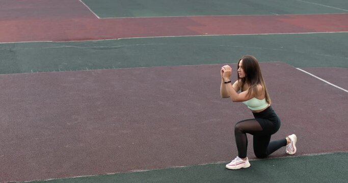 Athletic young woman is training, doing lunges on sport stadium ground in campus. Girl is in sportswear. Physical exercise for glutes and legs. Sport, fitness,wellness, workout outdoors concept.