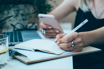 Woman's hand holding smartphone and making notes in notepad in cozy summer office with laptop
