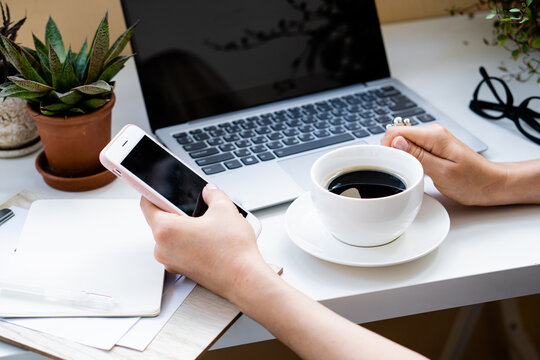 Woman's hand holding cup of coffee and smartphone in cozy office with laptop and green plants