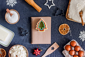 Cookbook with dark pages for writing a recipe surrounded by ingredients of flour, cottage cheese, butter, eggs, cookie cutters. Christmas cookie recipe. Top view, flat lay, copy space