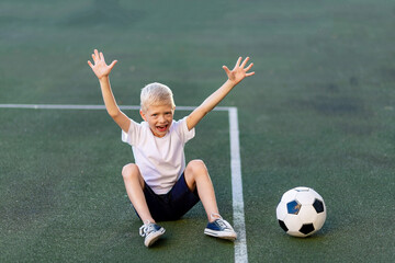 a blond boy in a sports uniform sits on a soccer ball on the football field and shouts Goal, sports section. Training of children, children's leisure