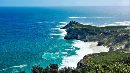 The Rocky Cape of Good Hope juts out into the ocean. Turquoise waves with white foam hit the stones. There is a small sandy beach between the mountains. South Africa.