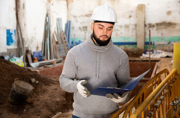 Focused young engineer planning renovation works at building site, looking at papers