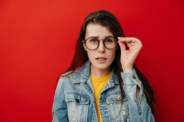 Shocked emotional young woman keeps hand on rim of spectacles, dressed in denim jacket, gazes with stupefaction, being amazed by terrifying news, isolated on red studio background. Reaction concept