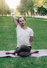 Fototapeta premium young man doing hatha yoga on green grass, lawn in city park in summer sunny time. A man sits on a rug, meditates, hands folded behind his back