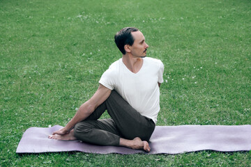 ..young man doing hatha yoga on green grass in city park in summer sunny time. Caucasian man sitting on mat in twisting sage asana