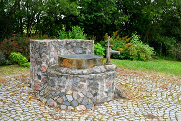 A close up on a well made out of small stones and boulders and with a brass tap situated in the middle of a public park next to some shrubs and a dense forest or moor spotted in Poland