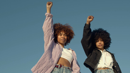 portrait of two mixed-race women with huge afro hair makes a protest black raised fist gesture outdoors portrait on the sky background - obrazy, fototapety, plakaty