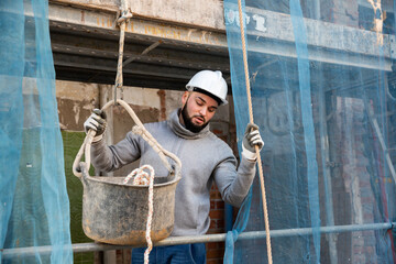 Confident bearded guy in construction helmet working on his house renovation, lowering down bucket with tools on rope..