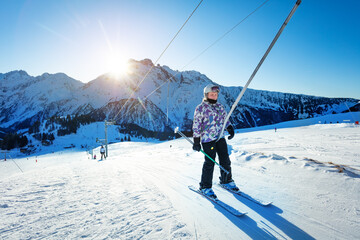 Happy teen girl lifting on the ski drag lift rope in bright sport outfit over sunset light near...