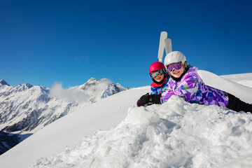 Two girls lay in the snow over blue sky and high mountains with ski on background