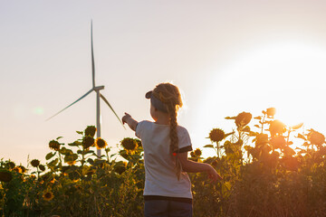 Cute girl in white t-shirt smelling sunflower in sunset field with wind turbines farm on...