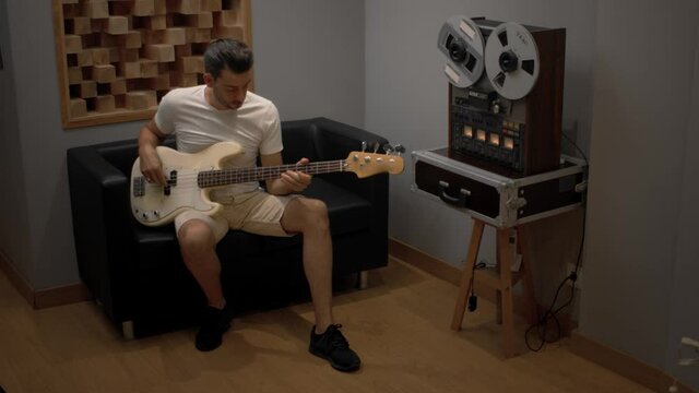 man playing bass guitar in sound studio with retro audio tape recorder at background