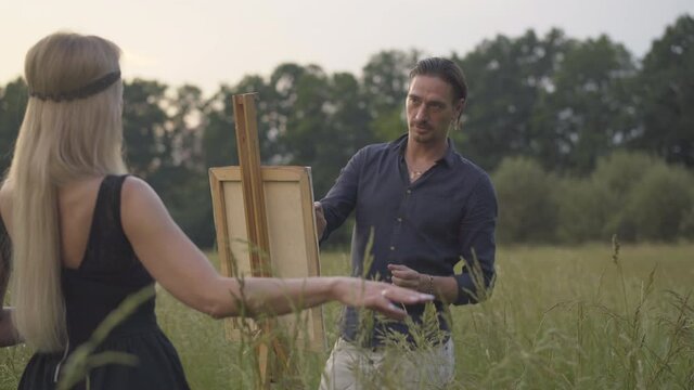 Back view of slim blond woman posing for artist painting picture outdoors. Concentrated handsome man drawing portrait of beautiful model at sunset on summer meadow.