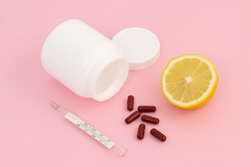 Brown pills near a white jar on a pink background, next to a thermometer and a slice of lemon.