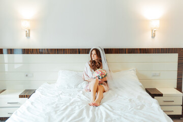 Morning of the bride in a pink robe and a veil in the hotel on the bed, wedding dress