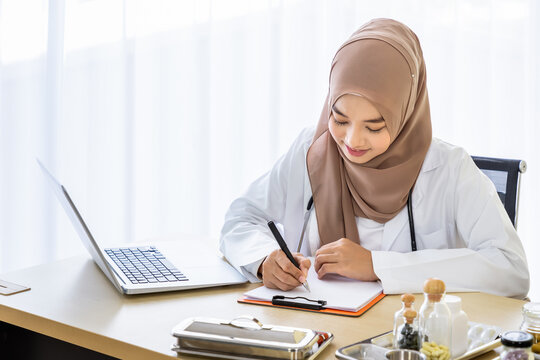 Confident Muslim female woman medical doctor sitting at desk, writing on notepad with laptop computer in clinic hospital office - Powered by Adobe