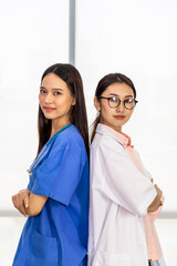 Two confident Asian female medical doctors standing back to back and cross arms; looking at camera