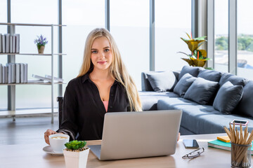 Portrait of young Caucasian woman with blonde hair freelancer working at home, sitting at desk in living room, looking at camera