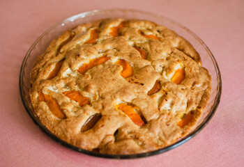 A large, round baked apple and apricot pie stands in a glass form on a table with a tablecloth.