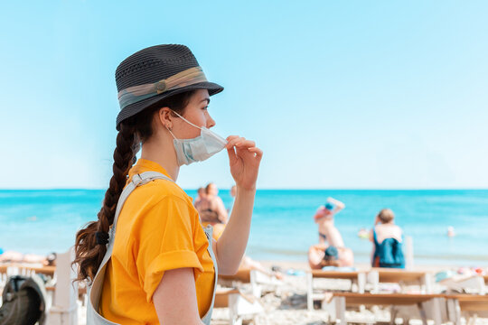 A woman removes the medical mask from her face to breathe in fresh air. In the background, the beach and the sea. The concept of vacation during a viral pandemic