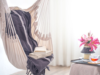 A boho-style hammock chair with a stack of books. Wooden tray with teapot and Cup of tea and beautiful Lily flowers. Cozy interior. The concept of rest and home comfort.