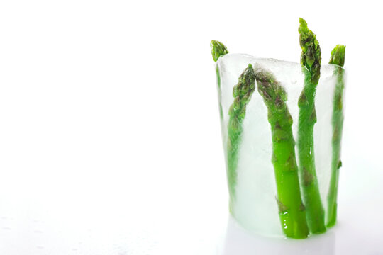 Cold transparent ice glass with frozen green asparagus inside on white background. Concept of frozen products. Space for text
