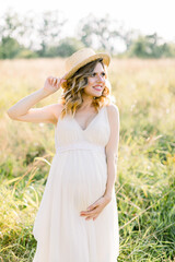 Fototapeta na wymiar Smiling charming young pregnant woman, wearing white dress and straw hat, resting in summer field. Posing outdoors. Motherhood. Outdoor portrait of beautiful pregnant woman in white dress