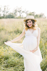 Outdoor portrait of young pregnant woman in the field. Pretty pregnant woman in white dress and hat relaxing in the summer nature meadow. Maternity concept