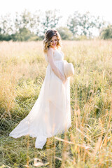 Fototapeta na wymiar Attractive young pregnant european woman in white dress, touching her big beautiful belly and walking in the meadow with sunset