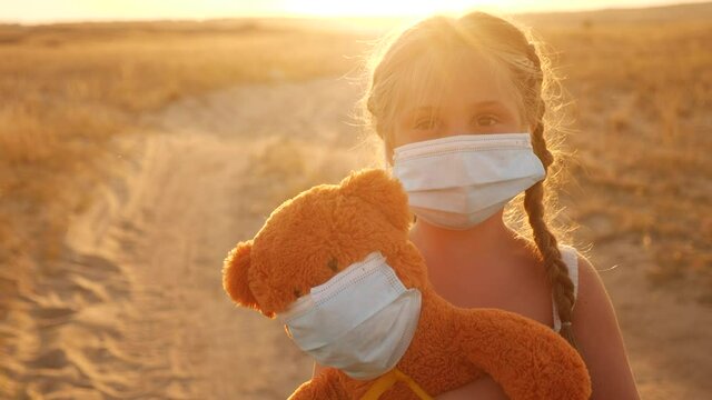 child girl in medical protective mask holding a teddy bear at sunset. concept pandemic coronavirus covid -19. girl kid face in a medical mask with a teddy bear toy. girl during coronavirus quarantine