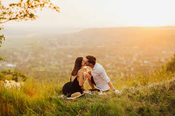 Fototapeta na wymiar Happy gorgeous elegant couple kissing on summer picnic. They are relaxing together on a summer day. Couple in love on a white plaid on mountain at a picnic against the background of the sunset