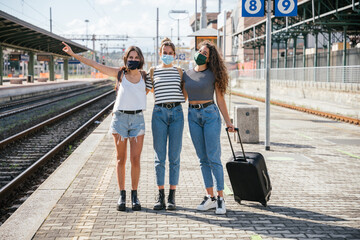 Three young friends women at the station waiting train for their trip in summer with face mask for...