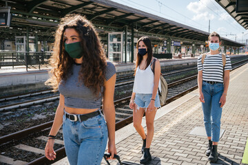 Three young friends women at the station waiting train for their trip in summer with face mask for...