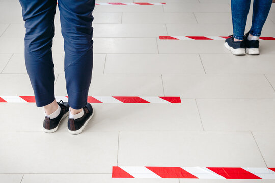 People Stand In Line Keeping Social Distance, Standing Behind Warning Line During Covid 19 Coronavirus Quarantine In Store Or Bank. Safe Shopping, Social Distancing Concept. Legs In Line Closeup