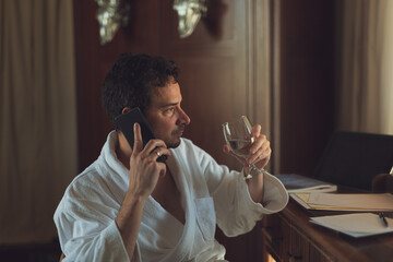 A man in a white bathrobe have a telephone call while holding a glass of water. During his stay at the desert resort.