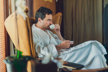 A man in a white bathrobe is having his first morning coffee at the hotel’s room seating on the...