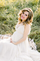 Fototapeta premium Outdoor summer photo of beautiful pregnant blond girl in white dress, sitting and resting on a checkered blanket in the summer sunny field