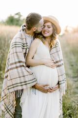 Handsome Caucasian man hugging his attractive pregnant woman in straw hat and white dress in sunny day in field. Happy couple covered with blanket, waiting for a baby