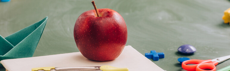 selective focus of delicious apple on book near school stationery on green chalkboard, panoramic concept
