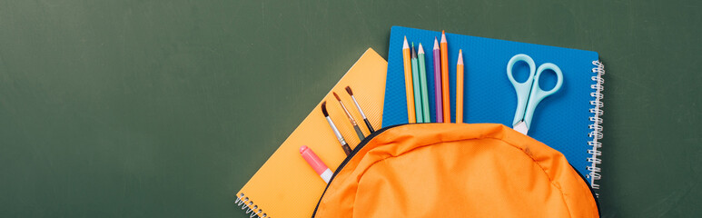 horizontal image of notebooks, color pencils, scissors and paintbrushes in yellow backpack on green chalkboard